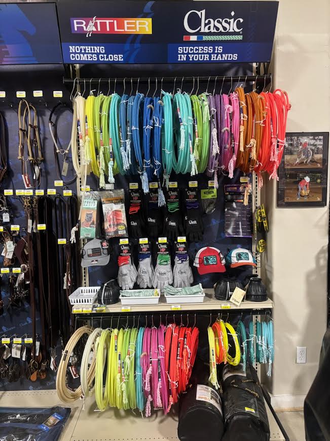 Western tack and roping supplies display in a store, featuring colorful rope coils, gloves, hats, and leather gear. The top signage highlights brands with slogans, while neatly organized products cater to rodeo and ranching needs.