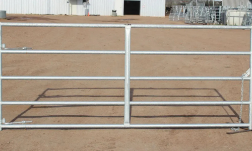 A metal farm gate with horizontal bars stands on a dirt path. In the background, there is a white industrial building, conveying a rural setting.