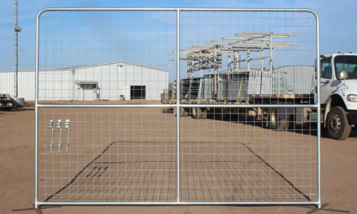 A large, double-panel metal gate stands on a dirt ground. Behind it, a white industrial building and a flatbed truck loaded with equipment are visible under a clear blue sky.
