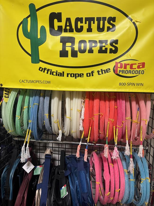 Store display featuring Cactus Ropes with a bright yellow banner, showcasing an assortment of colorful team roping ropes neatly arranged on a metal rack. Ropes in shades of green, blue, white, red, and pink are bundled with yellow ties, alongside rope bags below.